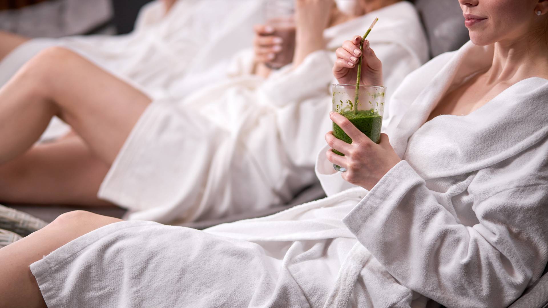 close-up-shot-young-women-wearing-white-bathrobes-drinking-freshly-squeezed-juice-while-spending-weekend-wellness-center-lying-bed-having-rest